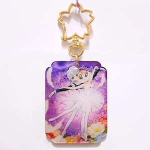May include: A rectangular keychain featuring an anime-style illustration of a ballet couple. The keychain has a gold-colored clasp and a flower-shaped top. The background is a mix of purple and floral patterns.