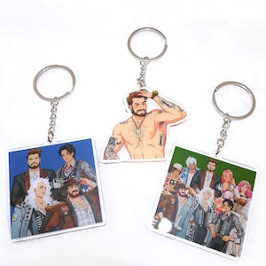 May include: Three keychains with character illustrations. One keychain features a shirtless man with tattoos, while the other two have group illustrations. Each keychain has a silver metal ring attached for keys or accessories.