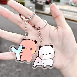 Pode incluir: Dois chaveiros com desenhos de ursos de desenho animado. Um urso é cor de pêssego segurando um "Y" azul, e o outro é branco com detalhes em preto segurando um "A" branco. Ambos os chaveiros têm argolas prateadas.