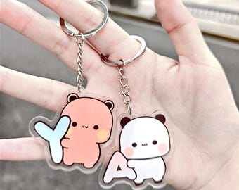 May include: Two keychains with cartoon bear designs. One bear is peach-colored holding a blue "Y", and the other is white with black accents holding a white "A". Both keychains have silver key rings.