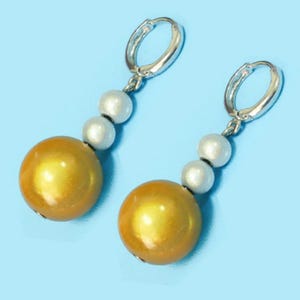 Könnte beinhalten: Ein Paar Ohrhänger mit silberfarbenen Creolen. Jeder Ohrring hat drei weiße Perlen und eine große, goldfarbene, kugelförmige Perle. Die Ohrringe sind vor einem hellblauen Hintergrund platziert.