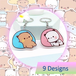 May include: Two acrylic keychains featuring cartoon bears. One bear is brown in a blue bed, the other is a panda in a pink bed. Each keychain has a silver chain and ring. The image also includes the text "9 Designs".