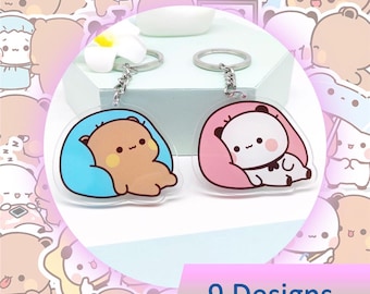 May include: Two acrylic keychains featuring cartoon bears. One bear is brown in a blue bed, the other is a panda in a pink bed. Each keychain has a silver chain and ring. The image also includes the text "9 Designs".