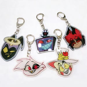 Puede incluir: Cinco llaveros acrílicos con diseños de personajes de dibujos animados. Los llaveros tienen anillos de metal plateado. Los personajes incluyen un demonio negro con un halo, un televisor con una cara de monstruo, un demonio pelirrojo, un personaje de pelo rosa y un personaje con sombrero.