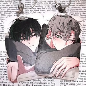 May include: Two anime-style acrylic keychains featuring male characters with black and gray hair, leaning on their arms. The keychains have silver-colored clasps and are set against a backdrop of printed text.