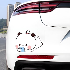 May include: A white car with a cartoon panda sticker on the back. The panda is white with black ears and brown spots, holding a blue phone. The panda is lying down, with a pink blush on its cheeks.
