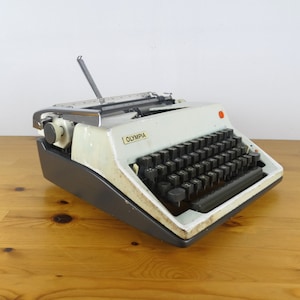 Puede incluir: Una m&aacute;quina de escribir Olympia vintage blanca con un teclado negro y un bot&oacute;n rojo en el lateral. La m&aacute;quina de escribir est&aacute; sobre una superficie de madera.
