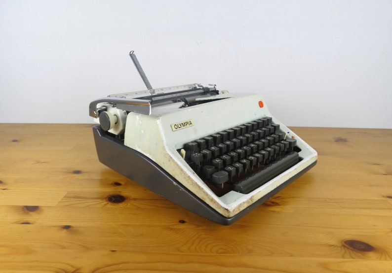 Puede incluir: Una m&aacute;quina de escribir Olympia vintage blanca con un teclado negro y un bot&oacute;n rojo en el lateral. La m&aacute;quina de escribir est&aacute; sobre una superficie de madera.