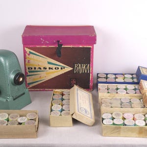 Puede incluir: Un proyector de diapositivas verde antiguo, una caja rosa con el texto "DIASKOP BAIKA" y varias cajas de diapositivas. Las cajas son de varios colores, incluyendo azul, rosa y beige. Los objetos están sobre una superficie blanca.