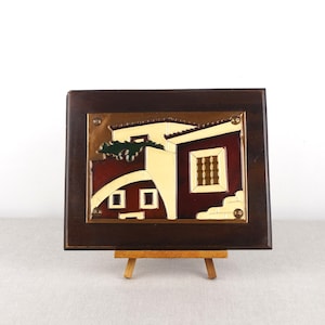 Puede incluir: Una obra de arte enmarcada que representa una ilustración estilizada de un edificio con un techo rojo, paredes blancas y un árbol verde. La obra de arte está montada en un caballete de madera.