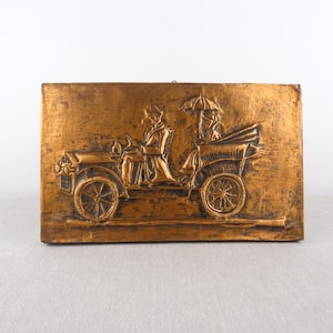 May include: A copper-colored wall plaque depicting a vintage car with two figures inside. The car is embossed with details and has a parasol above it.