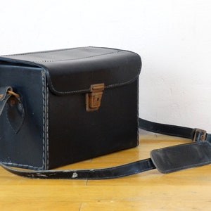 May include: A black leather camera case with a brown leather strap and a metal latch. The case has stitching around the edges.