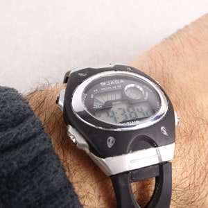 May include: A black and silver digital wristwatch with a black band. The watch face displays the time, date, and day of the week. The watch has a black and silver case with a silver bezel. The watch is on a person's wrist.