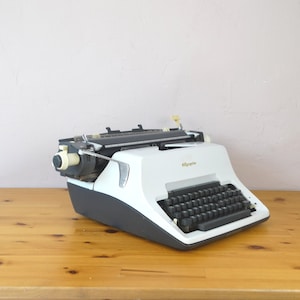 May include: A white and black vintage Olympia typewriter with a black keyboard and a white body. The typewriter is sitting on a wooden table.
