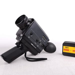 Può includere: Una cinepresa Super 8 nera vintage con maniglia e obiettivo, accanto a una cartuccia di pellicola nera con etichetta gialla. La fotocamera ha un cinturino nero e vari quadranti e pulsanti.