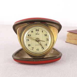 May include: A gold-toned alarm clock with a red leather case. The clock face has black Roman numeral hour markers and black hands. The clock is labeled "Star" and "7 Jewels".
