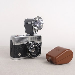 May include: A silver and black 35mm film camera with a flash attachment and a brown leather case. The camera has a lens labeled "Color-Apotar 1:2.8/40" and the text "Optima 200" on the front.