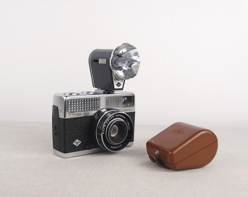 May include: A silver and black 35mm film camera with a flash attachment and a brown leather case. The camera has a lens labeled "Color-Apotar 1:2.8/40" and the text "Optima 200" on the front.
