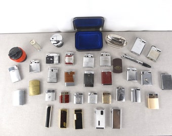 May include: A selection of vintage lighters in diverse designs and colours, including silver, gold, black, and red. Some lighters have textured finishes, while others are smooth. A pen and a small blue box are also visible.