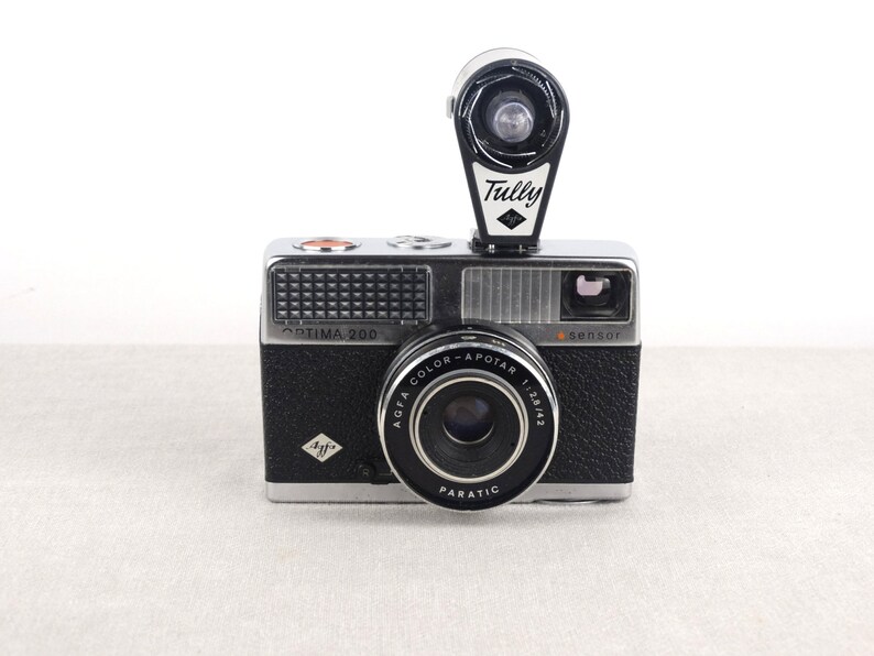May include: A black and silver Agfa Optima 200 camera with a built-in flash. The camera has a black lens with the text "Agfa Color-Apotar 1:2.8/43 Paratic" printed on it. The flash is a black and white Tully flash with a diamond logo.
