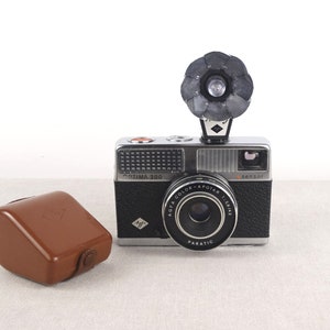 May include: A black and silver Agfa Optima 200 camera with a built-in flash and a brown leather case. The camera has a lens labeled "Agfa Color-Apotar 1:2.8/40" and "Paratic".