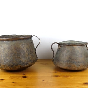 May include: Two antique copper pots with handles, one larger than the other, sitting on a wooden surface.
