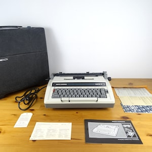 May include: A white Smith Corona electric typewriter with a black carrying case, instruction manuals, and a power cord. The typewriter has a black and gray keyboard and a black ribbon.