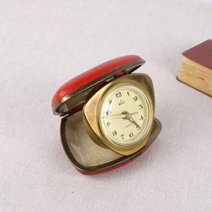 May include: A gold-colored alarm clock with a white face and black numbers. The clock is in a red leather case.
