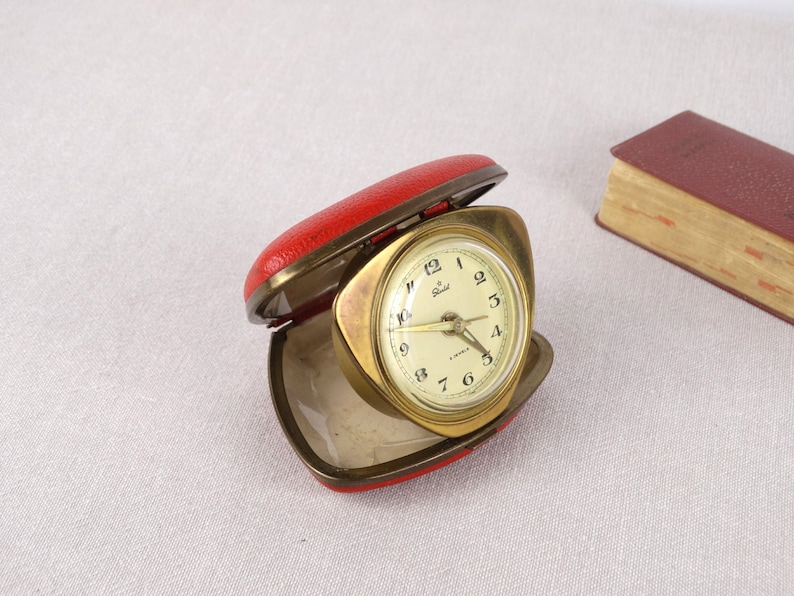May include: A gold-colored alarm clock with a white face and black numbers. The clock is in a red leather case.