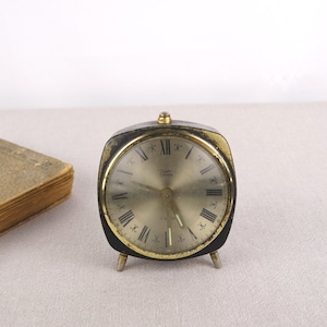 Könnte beinhalten: Ein vintage schwarzer und goldener Wecker mit römischen Ziffern auf dem Zifferblatt. Die Uhr hat eine quadratische Form und steht auf vier kurzen Beinen.