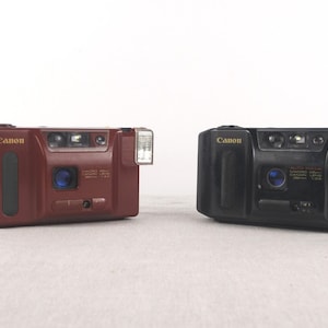 Puede incluir: Dos cámaras de película Canon Sure Shot de apuntar y disparar. Una es de color burdeos, la otra negra. Ambas tienen flash incorporado, una lente y el logotipo de Canon. La cámara burdeos tiene una empuñadura negra en el lateral. Las cámaras están sobre un fondo blanco.