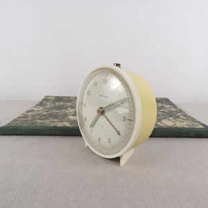 Puede incluir: Un despertador vintage crema con esfera blanca y números negros. El reloj tiene forma redondeada y una pequeña parte superior dorada. Se encuentra sobre un libro con estampado verde y gris. La esfera del reloj indica 'Junghans'.