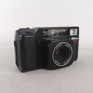 Pode incluir: Uma câmera Nikon Touch Zoom 300 preta. A câmera tem uma lente com o texto "Nikon LENS MACRO 38/65mm" e um flash embutido. A câmera está em uma superfície cinza claro.