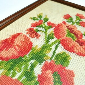 May include: A framed cross-stitch embroidery featuring a bouquet of pink flowers with green leaves. The flowers are arranged in a cluster and are surrounded by a white background.