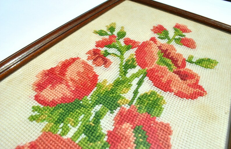 May include: A framed cross-stitch embroidery featuring a bouquet of pink flowers with green leaves. The flowers are arranged in a cluster and are surrounded by a white background.