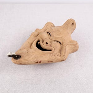 Può includere: Una maschera di legno scolpito color marrone chiaro con un volto sorridente, con la bocca spalancata e gli occhi chiusi. La maschera ha un piccolo foro sul lato con un filo bianco e nero attaccato.