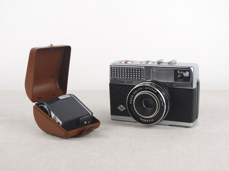 May include: A silver and black Agfa Optima 200 camera with a brown leather case. The camera has a lens labeled "Agfa Color-Apotar 1:2.8/45" and a flash attachment.