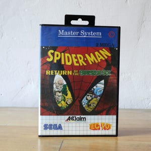 May include: A Sega Master System game cartridge for Spider-Man: Return of the Sinister Six. The cartridge is black with a blue and red label featuring a close-up of Spider-Man's face and the game title. The label also includes the text "2 MEGA" and "Aklaim".
