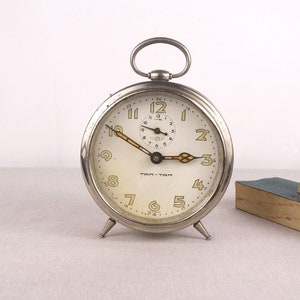 May include: A silver vintage alarm clock with black hands and Roman numeral markings on the face. The clock has a white face with black numbers and the words "Tom-Tom" printed on it. The clock is sitting on a white surface.