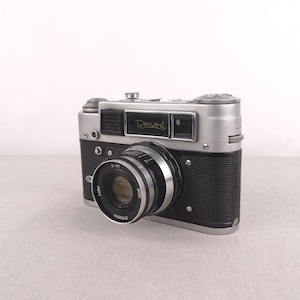 Puede incluir: Una cámara de película de 35 mm de color plateado y negro con el texto "Revue-4" en la parte superior. La cámara tiene una cubierta de cuero negro en la parte inferior y una lente plateada con el texto "Jupiter-8" en ella.
