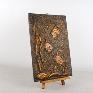 Puede incluir: Un colgante de pared de madera con una superficie texturizada y tres peces tallados en relieve. Los peces son de color dorado claro y el fondo es marrón oscuro. El colgante de pared está sobre un pequeño caballete de madera.