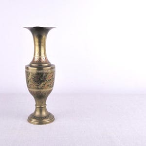May include: A brass vase with an ornate floral design. The vase has a flared top and a pedestal base. The body of the vase is decorated with etched patterns and a band of reddish-brown accents. The vase is a warm gold color.