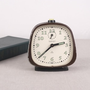Puede incluir: Un reloj despertador vintage marrón y crema con el nombre "peter" en la esfera. El reloj tiene una base negra y una esfera crema con números y manecillas negras. El reloj está sobre un fondo neutro.
