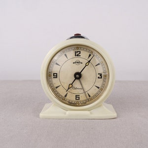 May include: A white vintage alarm clock with black hands and a cream-colored face. The clock face has black Roman numeral hour markers and black Arabic numeral minute markers. The clock face also has the word "Сделано" in Cyrillic script.
