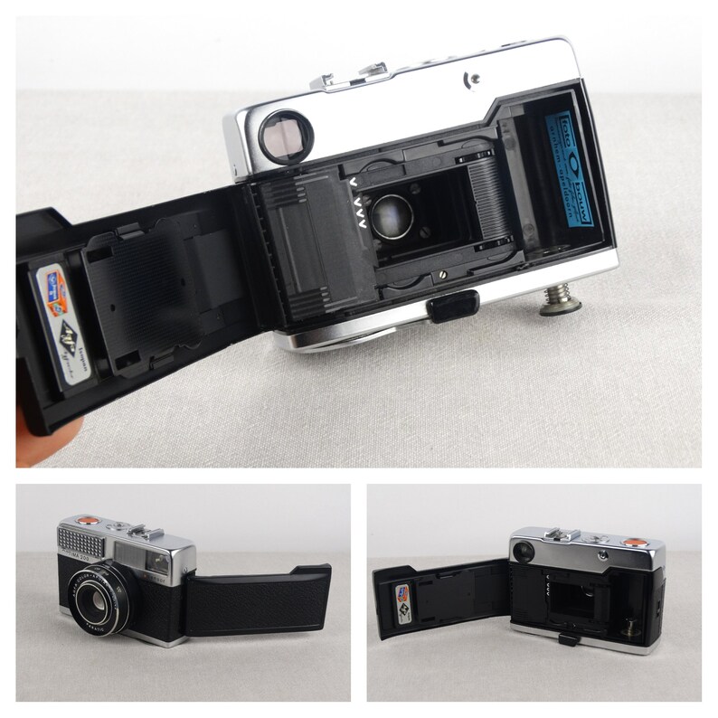 May include: A silver and black 35mm film camera with a built-in flash. The camera has a black leatherette covering and a silver lens. The camera is open, showing the film compartment.