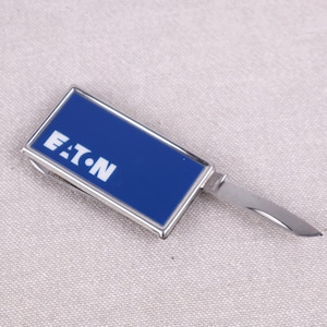 Puede incluir: Una pequeña navaja de bolsillo rectangular con un diseño azul y plateado. La sección azul presenta el texto blanco "EATON". La hoja del cuchillo está extendida. El artículo está sobre un fondo neutro.