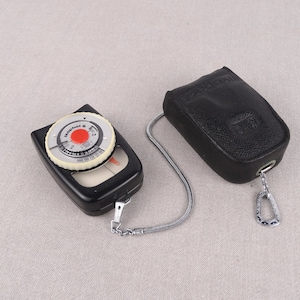May include: A black light meter with a white dial and a red needle. The light meter is attached to a black leather case with a silver chain.