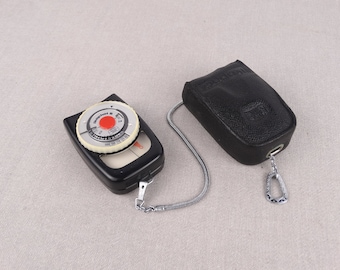 May include: A black light meter with a white dial and a red needle. The light meter is attached to a black leather case with a silver chain.