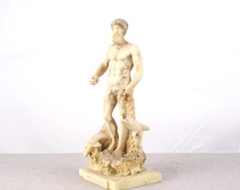 May include: A white marble statue of a nude man standing on a rocky base with two dolphins. The statue is about 12 inches tall and is made of a white marble-like material.