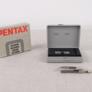Può includere: Una custodia in plastica grigia con un portapellicola in metallo argentato e un clip in metallo argentato per pellicola. La custodia ha la scritta "PENTAX" stampata in lettere rosse. La custodia è etichettata "Accessory, Accessoire, Zubehör, Accessorio".
